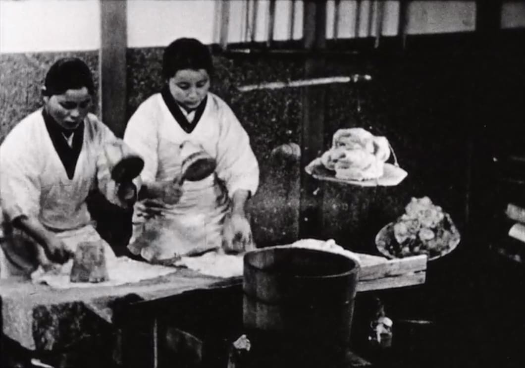 Two women work side by side at a table, pounding tree bark with wooden mallets as part of a traditional processing method class=