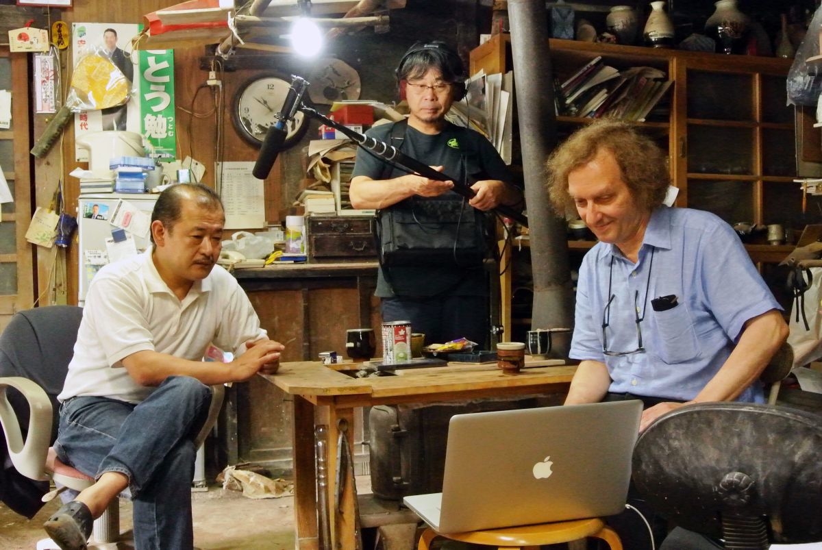 Marty Gross reviewing footage on a laptop while interacting with a seated man across the table, as a sound operator holds a boom microphone in the background