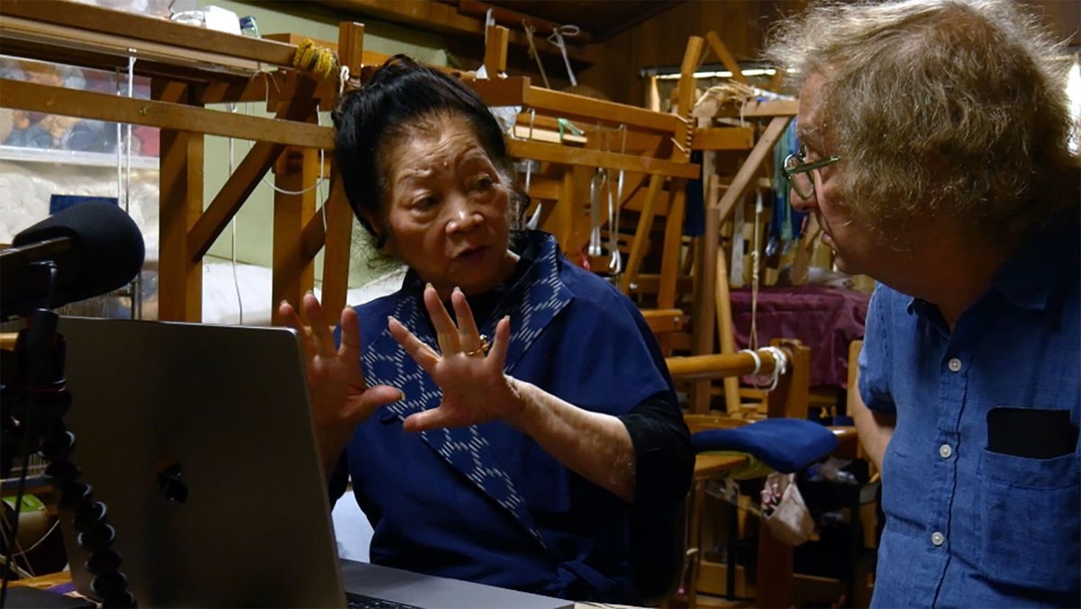 Inside a weaving workshop, a female artisan gestures as she explains something while Marty Gross listens attentively across from her