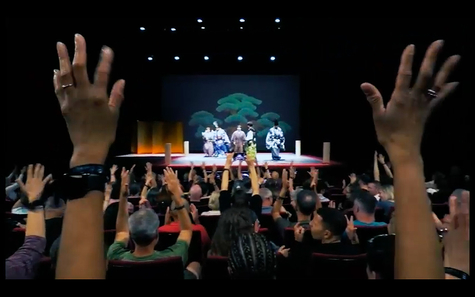 A scene from the Paris performance held at the Japan Cultural Institute in Paris. On stage, performers are wearing traditional Japanese costumes, with a green pine tree painted in the background. Many people are seated in the audience, raising their hands in applause