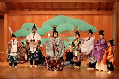 A scene from a Sign Language Kyogen stage performance. Performers in traditional Japanese costumes are performing on a stage with a green pine tree painted in the background