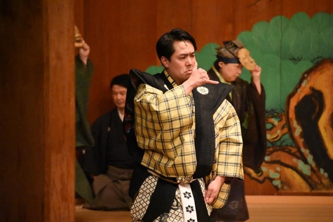 A scene from a Sign Language Kyogen stage performance. A performer in a traditional Japanese costume with a black and yellow checkered pattern stands on stage, holding his hand near his face