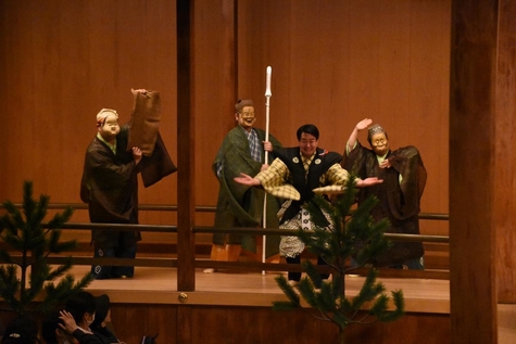 A scene from a Sign Language Kyogen stage performance. Four performers are standing on stage, three are wearing masks, and the remaining one is performing without a mask