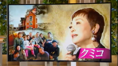 A scene from the program is displayed on a large monitor. On the right half of the monitor is a caption that reads 'Kumiko,' along with a profile of a woman holding a microphone. On the right half is a burning building that was bombed, and underneath that are photos of a dozen family members.