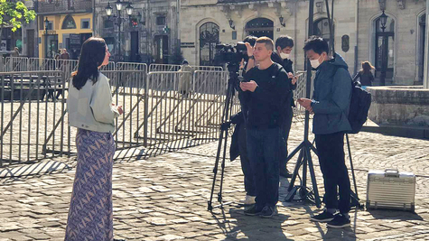 An interview on the street. A woman in a white jacket stands on the left side of the screen, and several male staff members are conducting an interview with a camera on a tripod on the right.