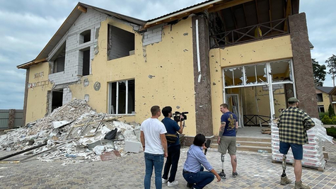 Three male staff members, one of whom is holding a handheld camera, are conducting an interview in front of a damaged house. Two men with prosthetic left legs from the thigh down are standing there, appearing to be being interviewed.