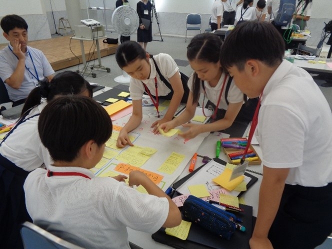 机を囲んで議論し合う制服姿の小学生たちとそれを見守る大人