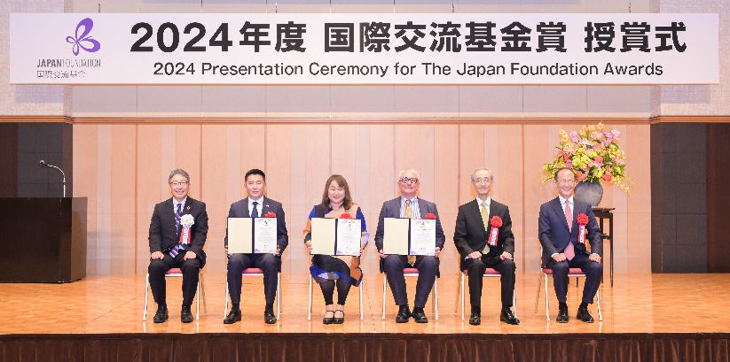 壇上に5人の人物が5脚のいすを並べて座っており、中央の3人の受賞者が盾を持っている受賞風景