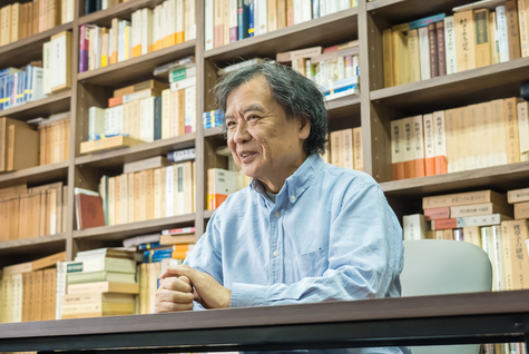 本棚の前でインタビューを受ける片渕監督　斜め右下の角度から