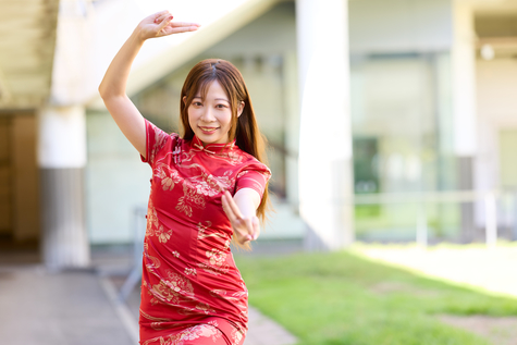 屋外で半袖の赤いチャイナドレス姿の女性がこちらを向いて右手を上に、左手を前に突き出してこちらを向いて微笑んでいる